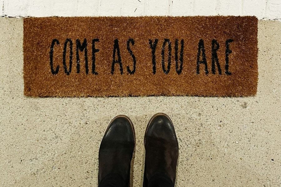 A coir doormat reading Come As You Are, seen from above with someone's boots standing in front of it.